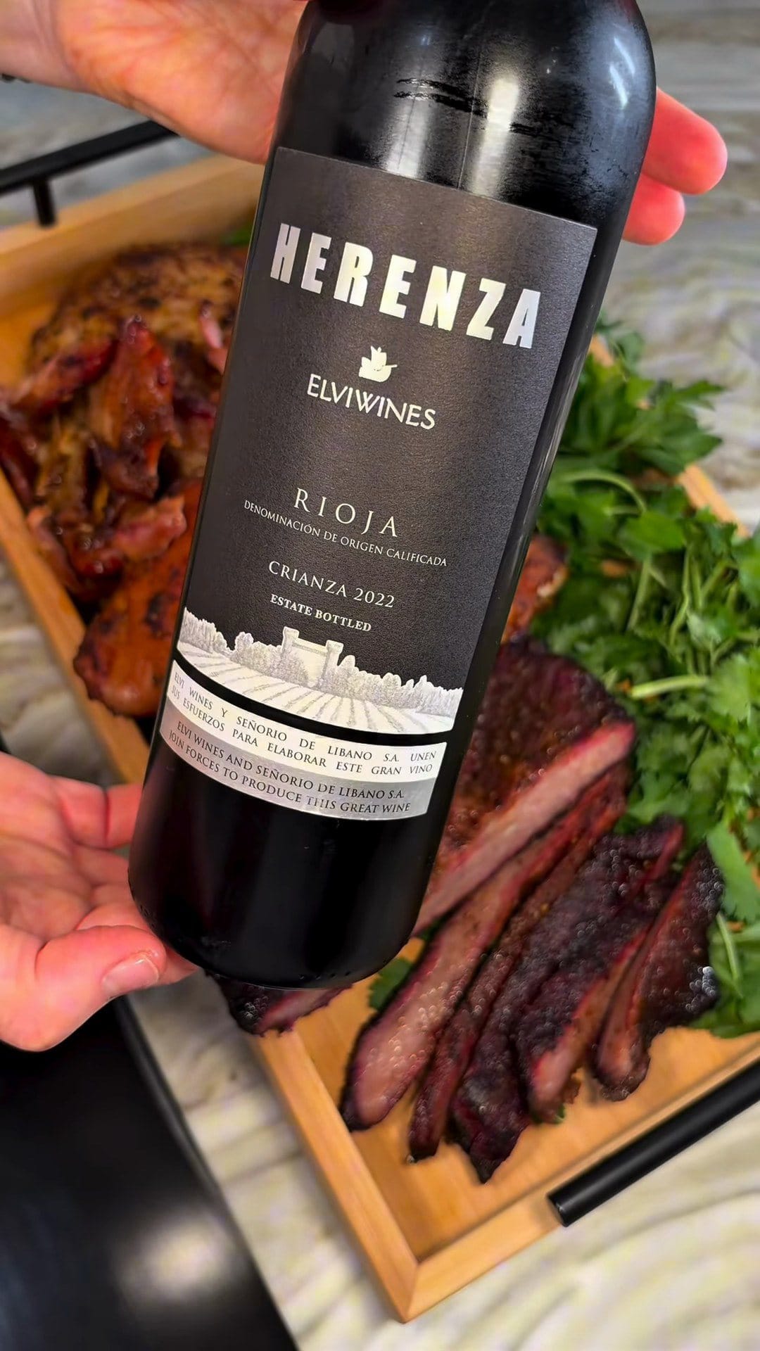 Bottle of Herenza Crianza 2022 by Elvi Wines, Rioja, held up in front of sliced smoked brisket, chicken and cilantro on a wooden tray