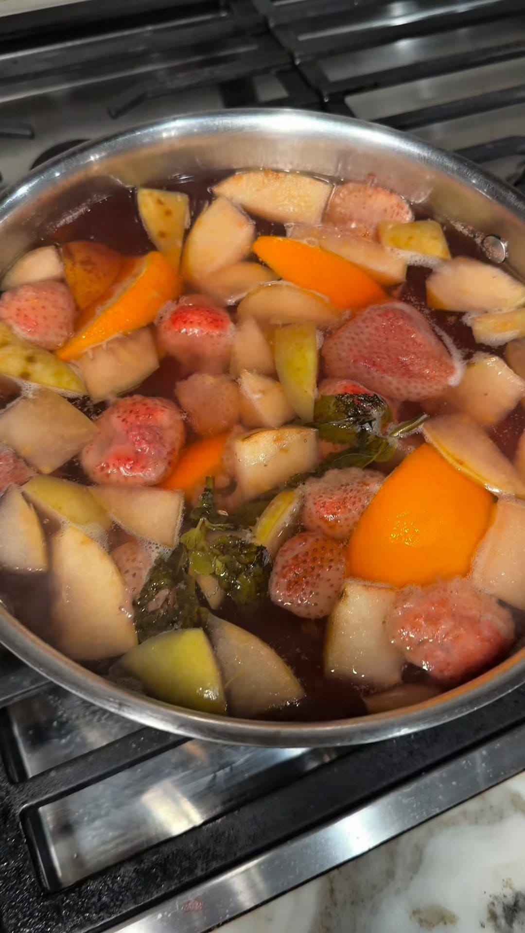 Apples, pears, strawberries, orange wedges and fresh mint simmering in a pot of rosy liquid on the stove