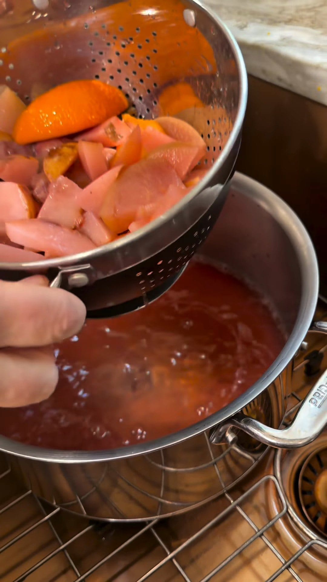 A colander full of simmered apple, pear and orange pieces lifted over a pot of rosy strained kompot