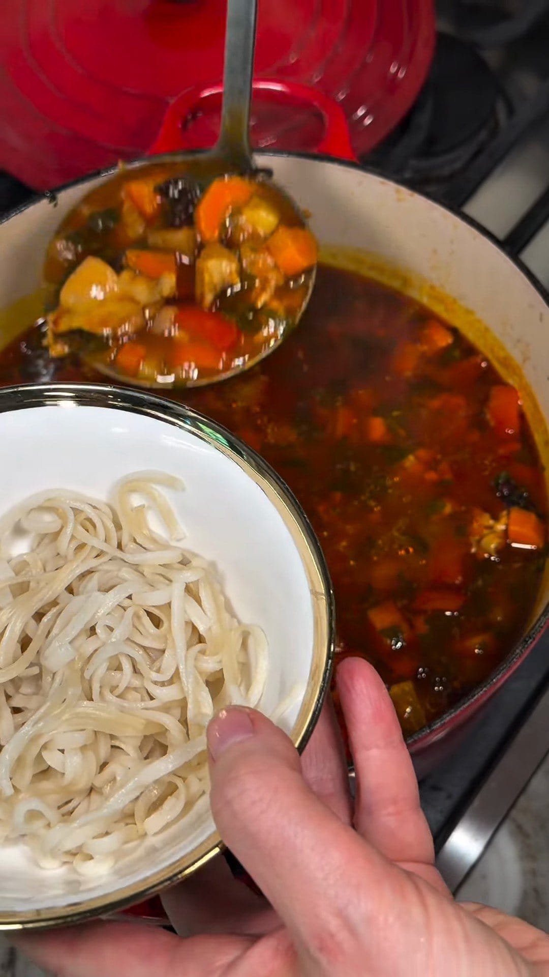 A bowl of thick white noodles held beside the pot as a ladle lifts out chunky stew