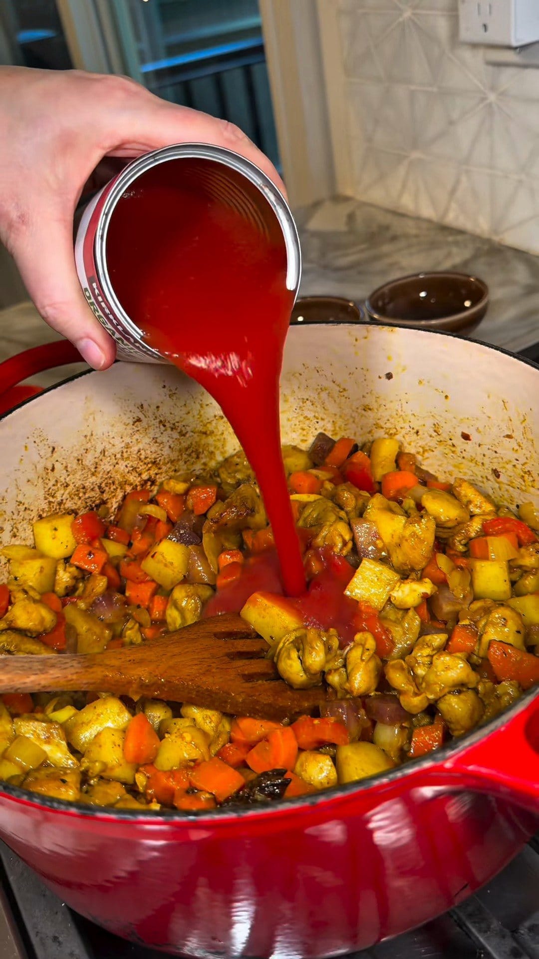 Thick tomato sauce pouring from a can over the chicken and vegetables in the red Dutch oven