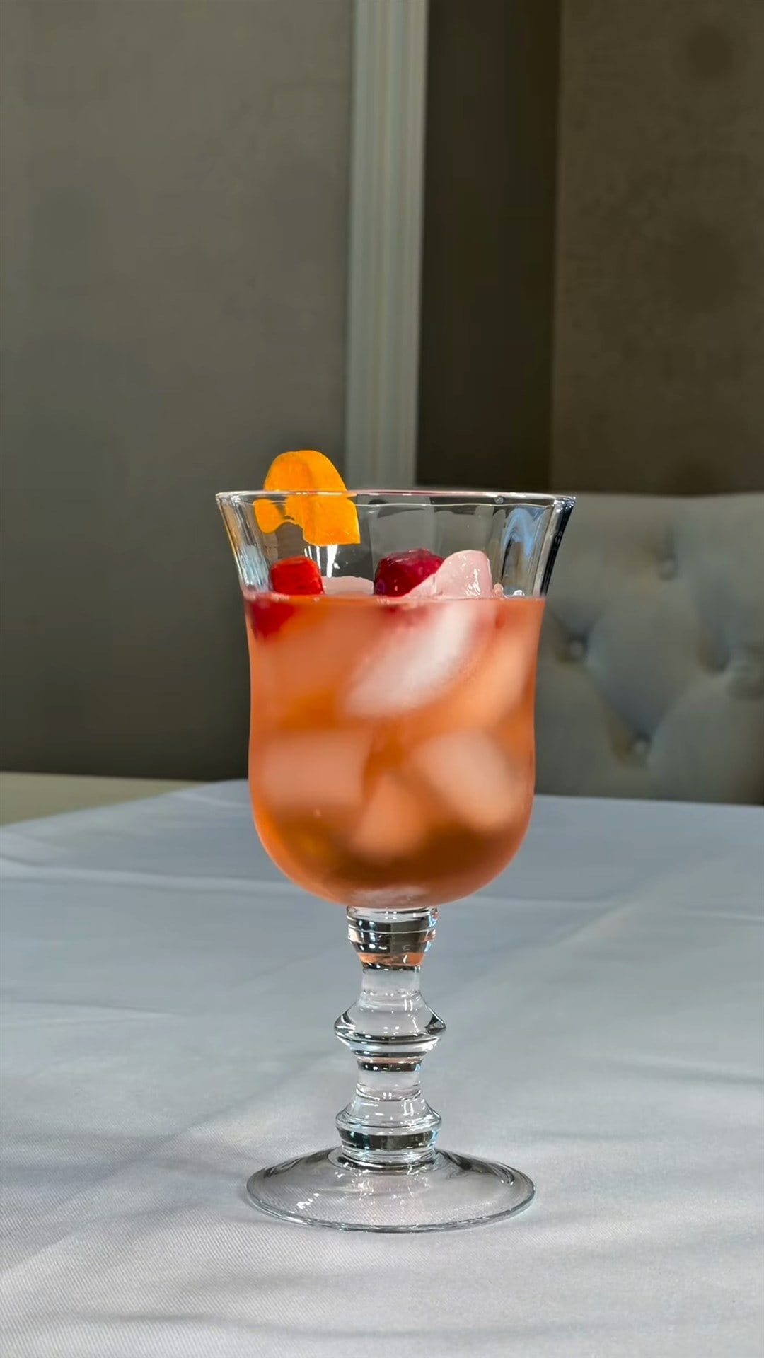 A stemmed glass of rosy fruit punch over ice, topped with strawberries and an orange-peel twist, on a white tablecloth