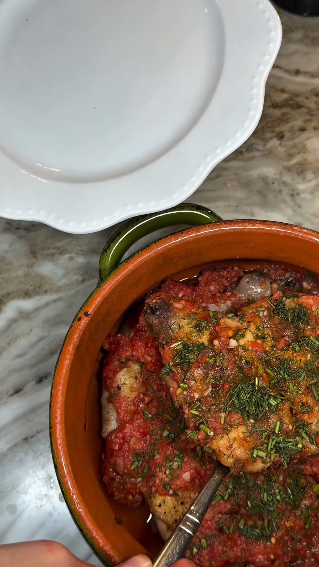 Overhead view of crispy chicken under fresh tomato sauce, chopped dill and chives in a terracotta dish, beside an empty white plate