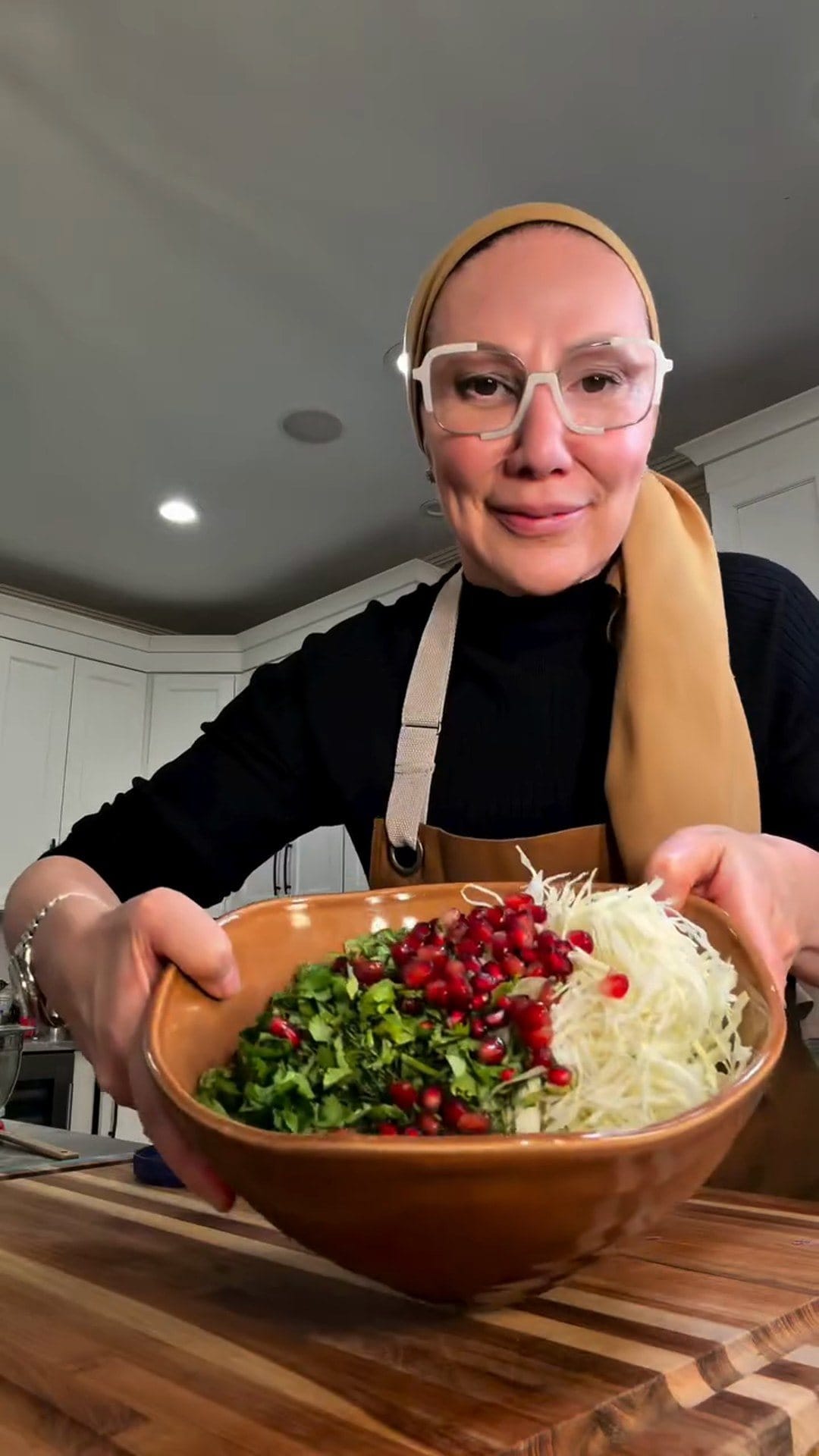 Anne in a tan apron and headscarf holding up a brown bowl of shredded white cabbage, chopped green herbs and pomegranate seeds in her kitchen