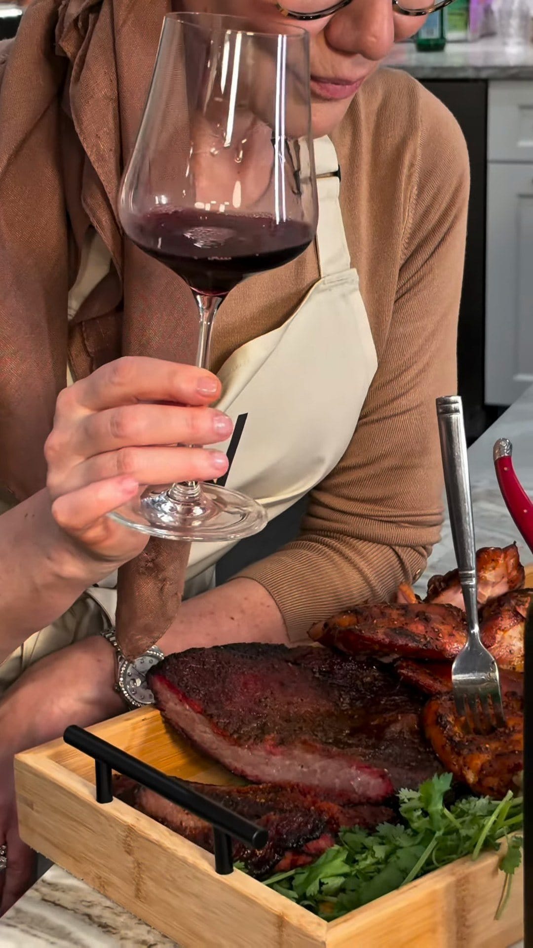 Anne sniffing a glass of Herenza Crianza over a wooden tray of sliced smoked brisket, chicken thighs and cilantro
