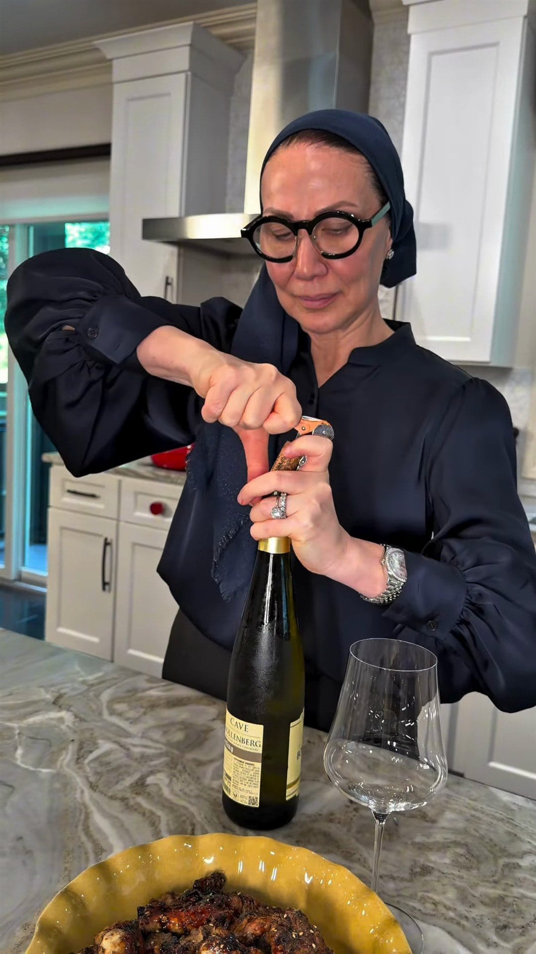 Anne at her kitchen counter uncorking a bottle of Cave du Bollenberg Riesling, an empty wine glass and the yellow bowl of wings in front of her