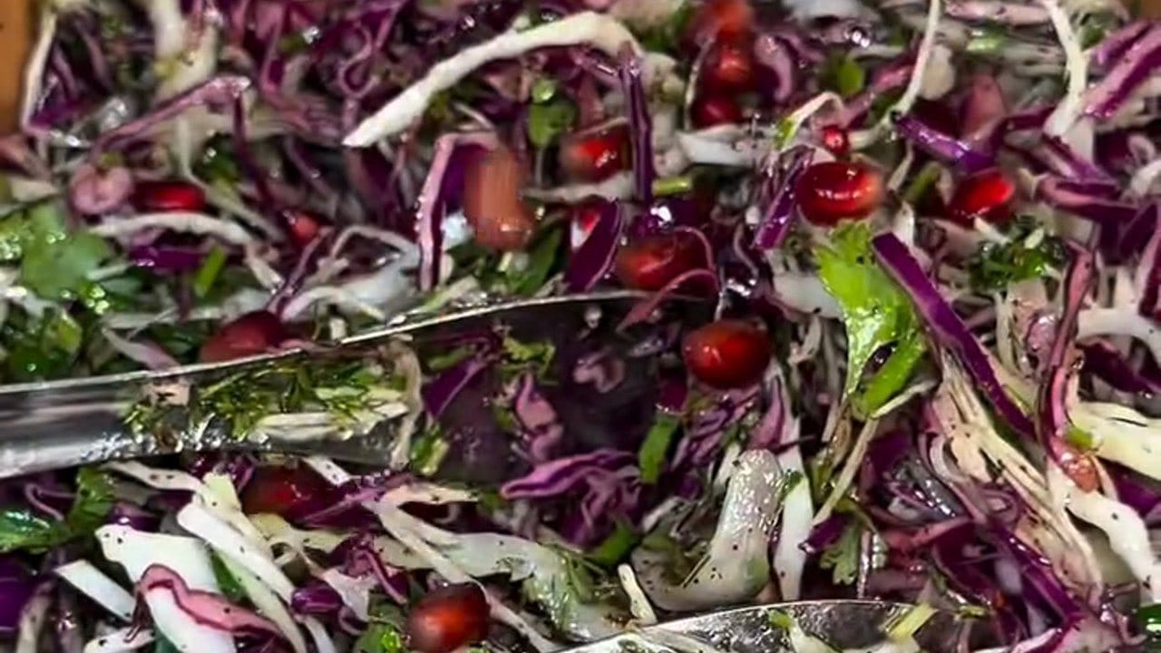 Close-up of the tossed salad: shredded red and white cabbage, green herbs and pomegranate seeds glossy with dressing, with silver serving tongs