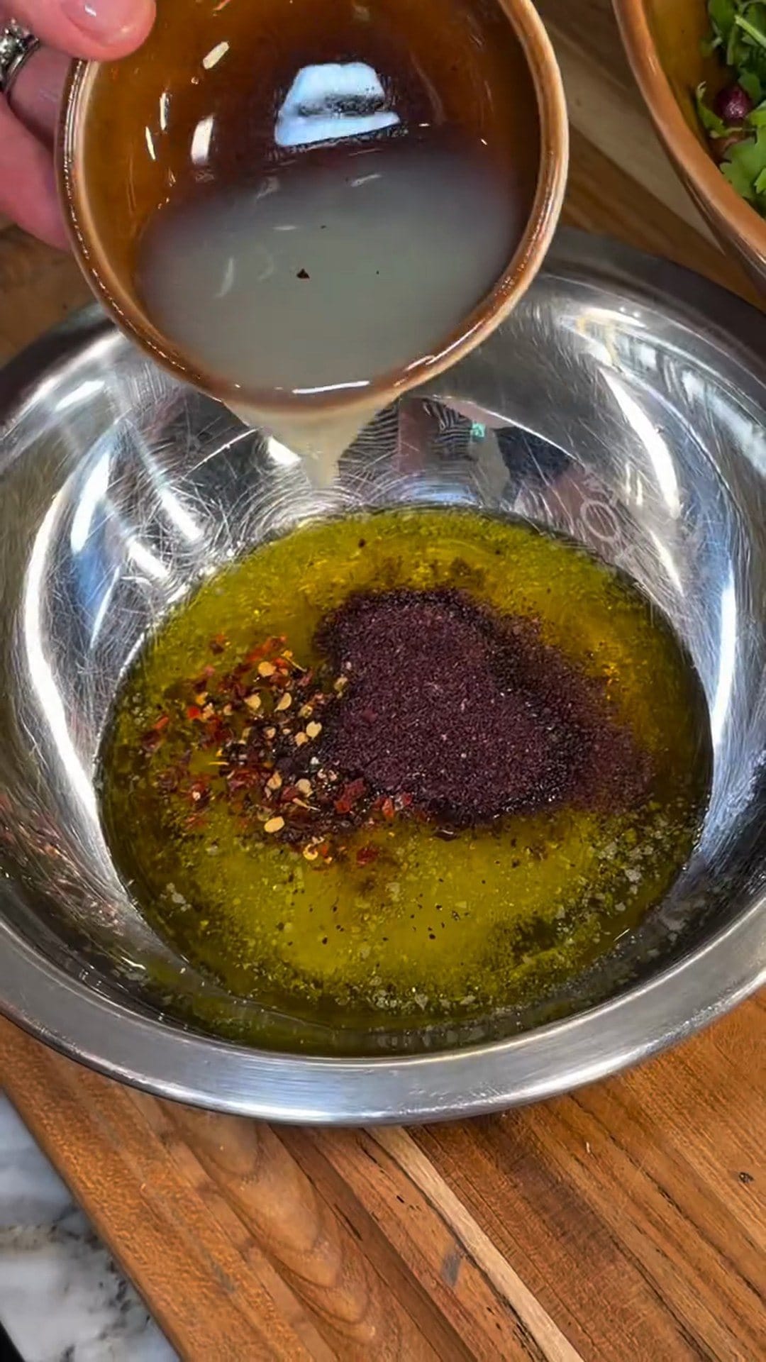 Fresh lemon juice pouring from a small brown bowl into olive oil topped with a mound of dark red sumac and red pepper flakes in a steel bowl