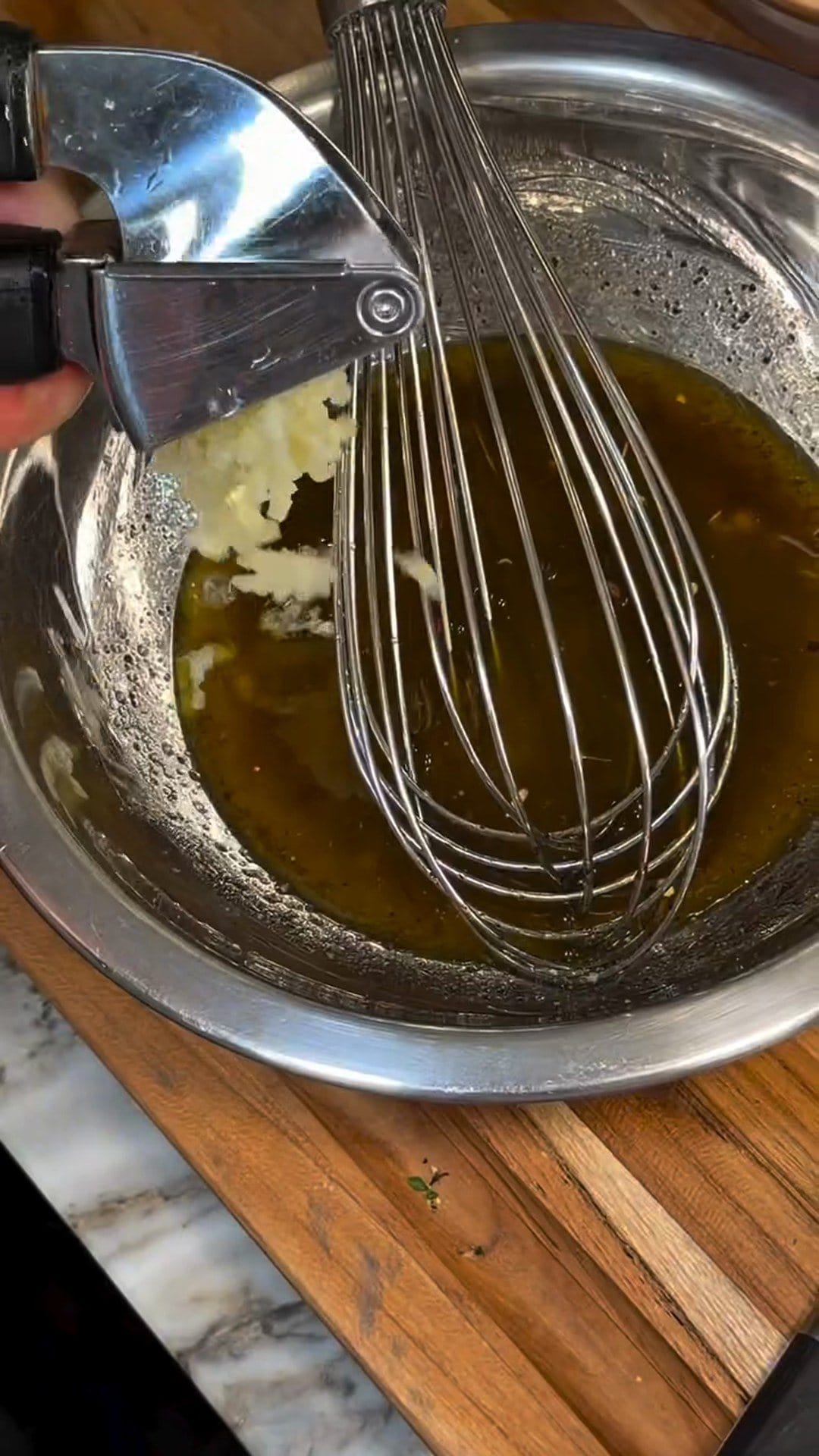 Crushed garlic coming out of a garlic press into the whisked lemon and olive oil dressing, a balloon whisk in the steel bowl