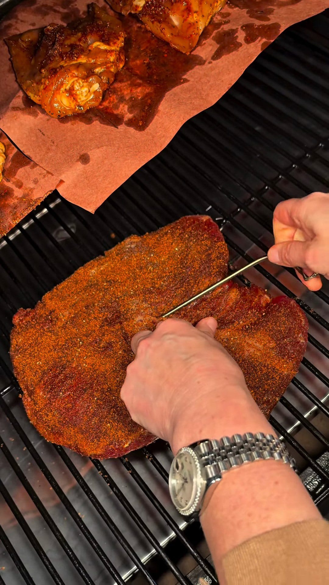 The rubbed brisket on the smoker grates with a temperature probe going in, chicken thighs on butcher paper beside it