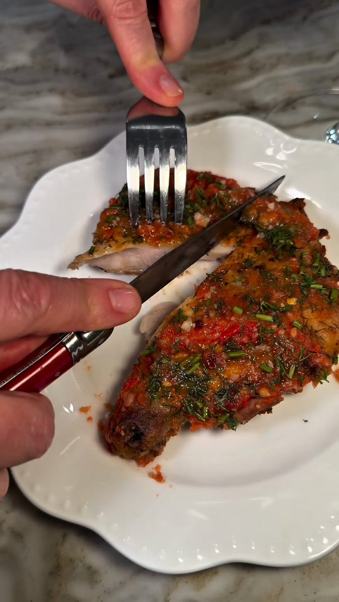 A knife and fork cutting into a crispy chicken thigh topped with tomato, garlic, dill and chives on a white scalloped plate, showing the juicy meat inside
