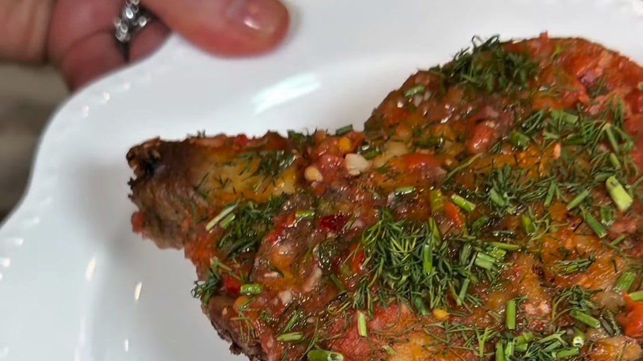 Close-up of a crispy chicken thigh on a white plate, covered in tomato, minced garlic, chopped dill and chives