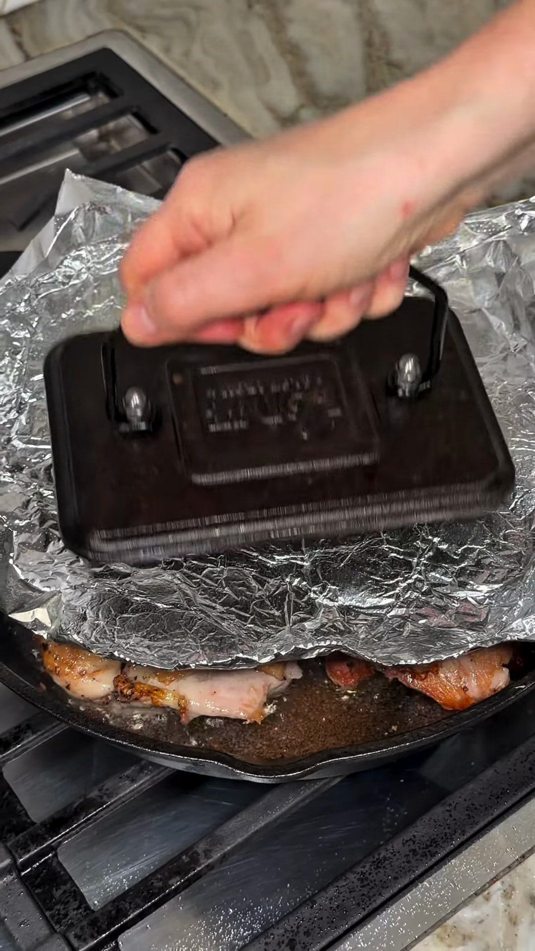 A cast-iron grill press set on aluminum foil over chicken searing in a cast-iron skillet