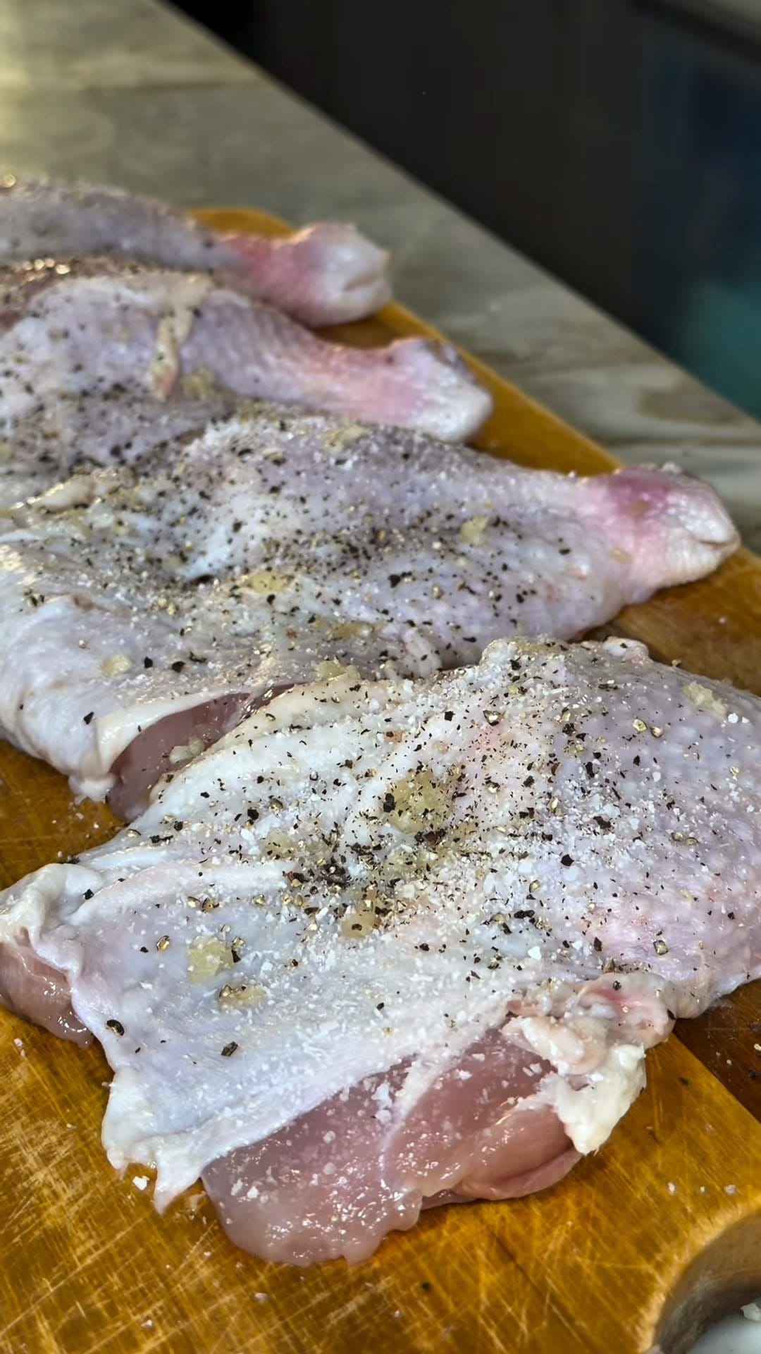 Boneless skin-on chicken thighs on a wooden board, sprinkled with salt and cracked black pepper