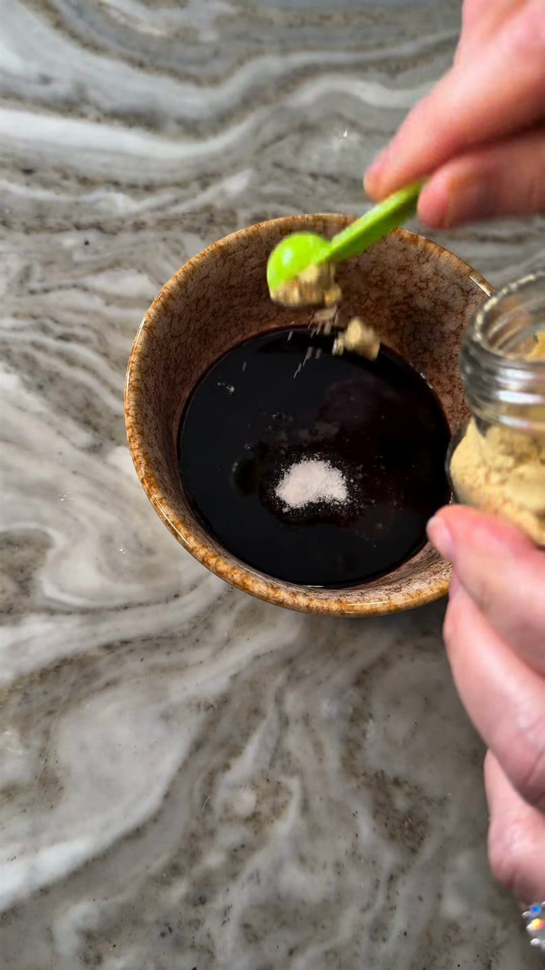A green measuring spoon adding seasoning to dark tamarind glaze in a small speckled bowl