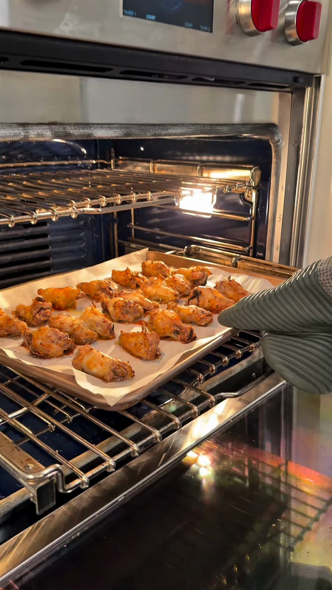An oven-mitted hand sliding a parchment-lined sheet pan of golden roasted wings into the oven
