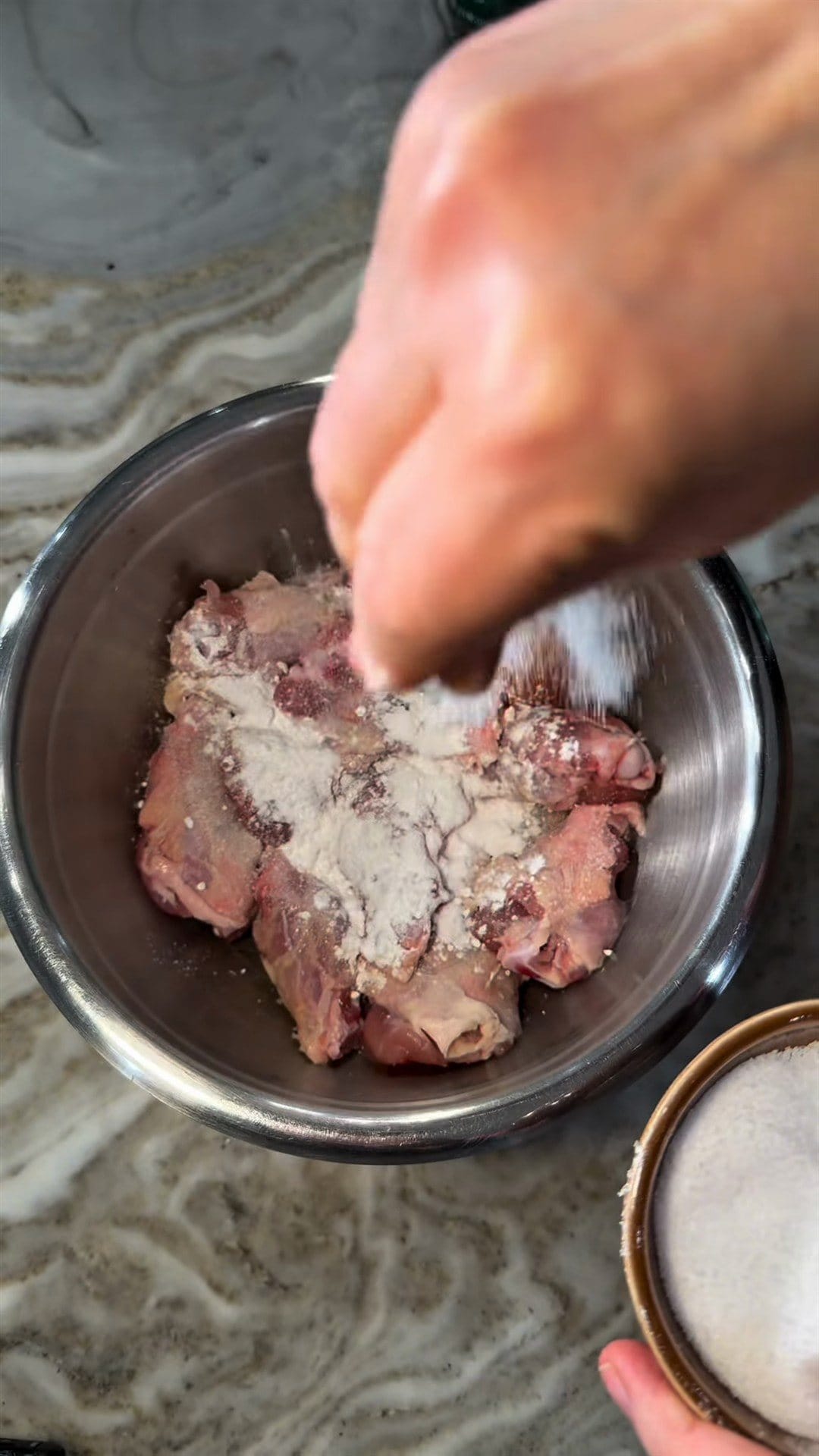 Anne's hand sprinkling baking powder and seasoning over raw chicken wings in a steel bowl, a dish of kosher salt beside it