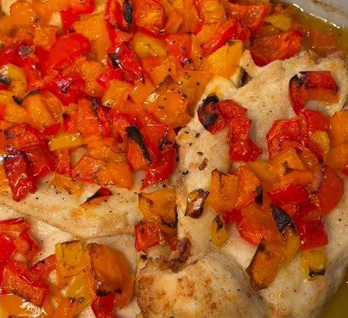 Close-up of baked chicken cutlets under a heap of roasted red, orange and yellow bell peppers in olive oil