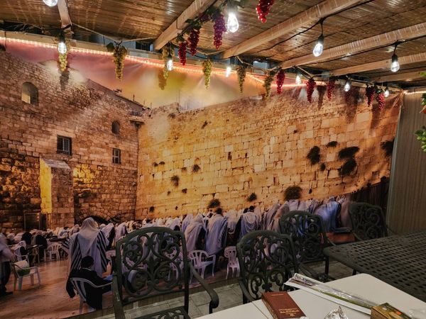 Inside a sukkah at night: grape clusters and string lights hang from the schach above a table, with a mural of the Western Wall behind it