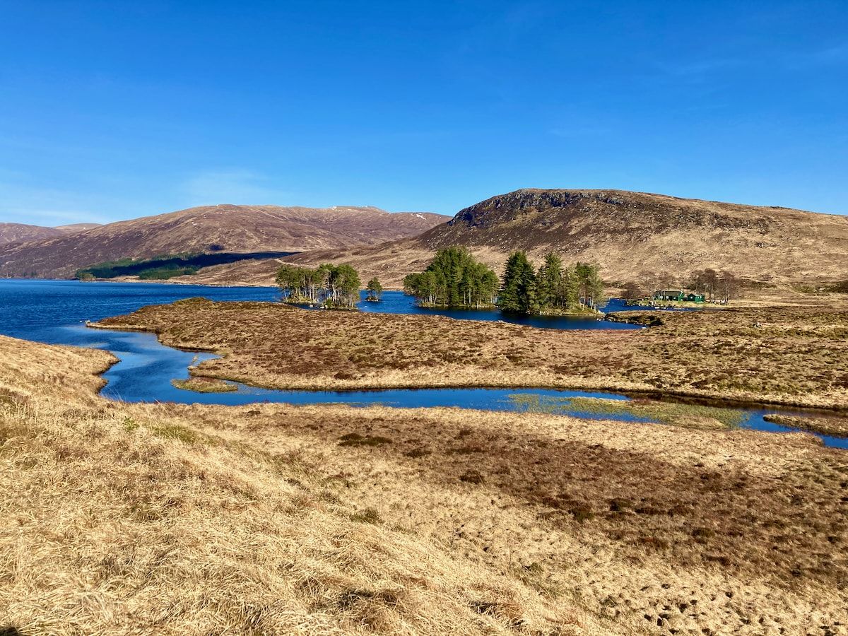 Loch Ossian mit kleinen Inseln mit Bäumen, rechts im Hintergrund ein Haus