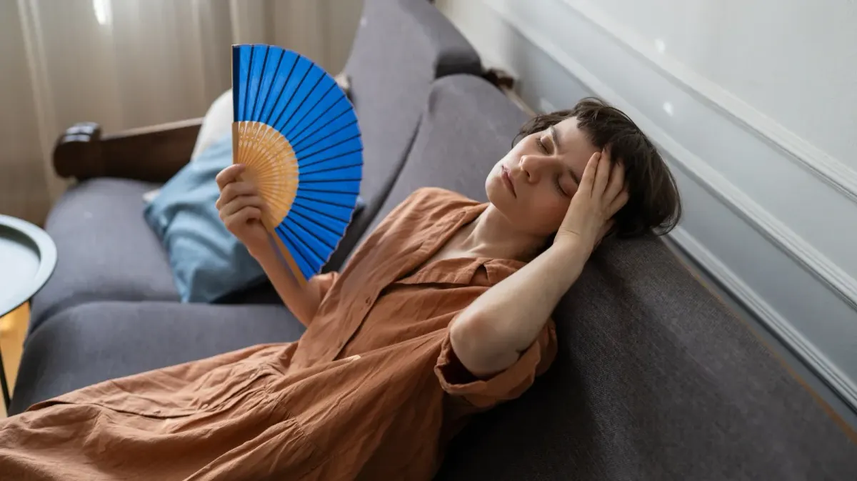 Woman sitting indoors looking overheated and fatigued during hot weather while cooling herself with a hand fan