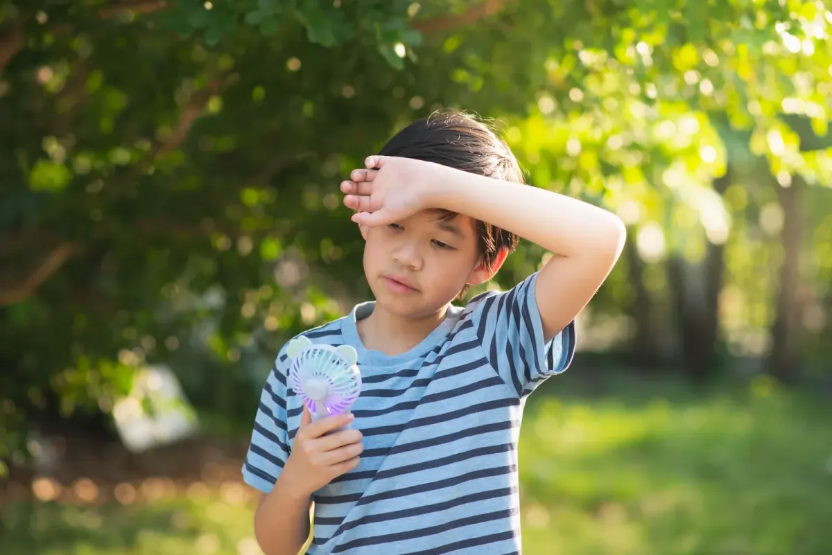 Child appearing overheated outdoors while cooling down with a handheld fan during hot weather