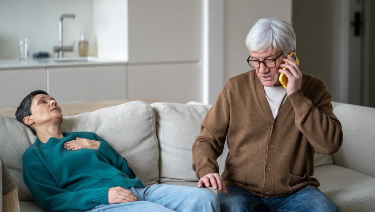 Older man calls emergency services while a woman lies back on a sofa feeling unwell.