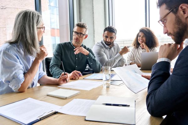 Employees in a workplace meeting discussing documents and reports around a conference table.