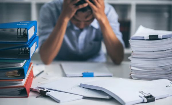 Office worker holding head at desk surrounded by paperwork, representing workplace stress and overload