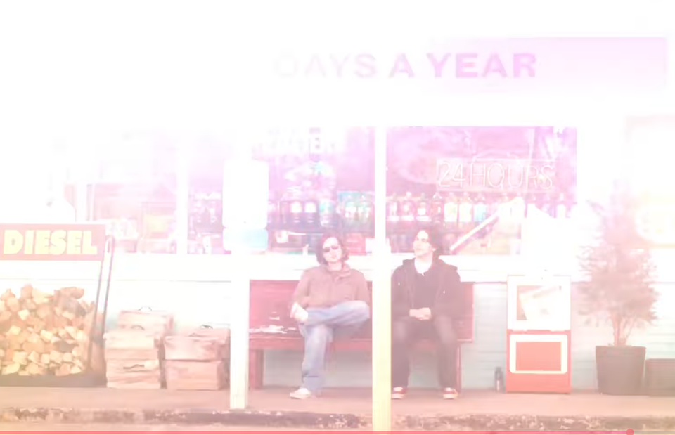 Screenshot showing two people sitting on a bench in front of a store from The Great Divide music video.