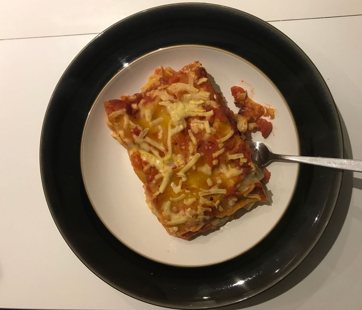 A slice of tasty, PKU Lasagne plated on a white plate with a wide black rim. A fork is to the side, and the plate rests on a white background