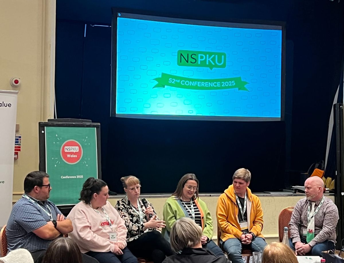 Photo of 6 people holding a discussion under two presentation boards. Reading NSPKU 52nd Conference 2025 and the other reading NSPKU Wales