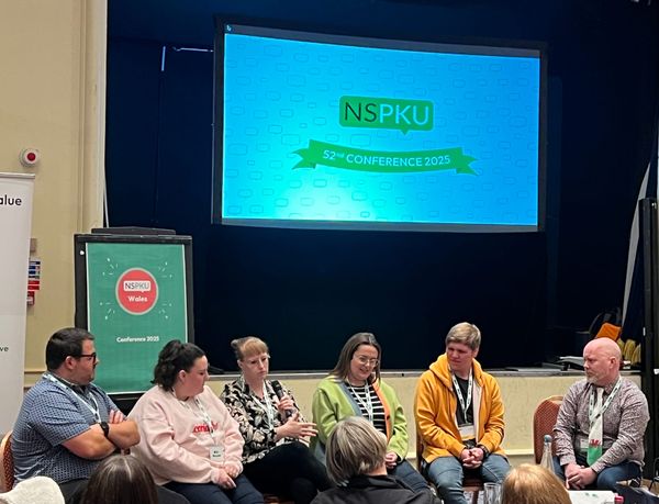 Photo of 6 people holding a discussion under two presentation boards. Reading NSPKU 52nd Conference 2025 and the other reading NSPKU Wales
