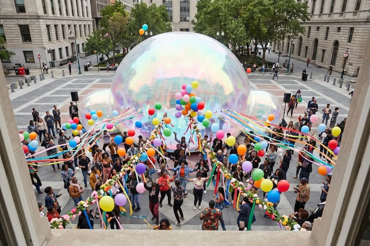 Grote iriserende koepel op openbaar plein, omringd door mensen met kleurrijke ballonnen en linten