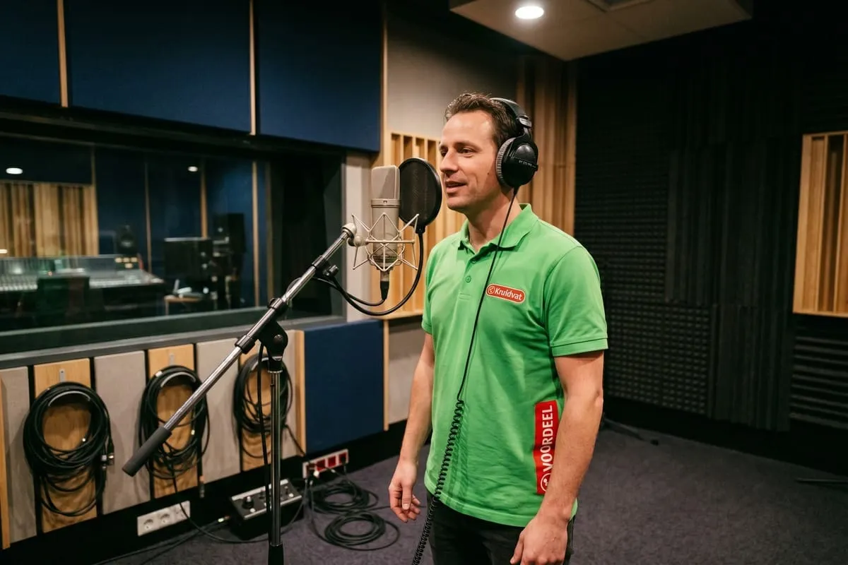 Man in groen Kruidvat-polo neemt audio op achter studiomicrofoon met koptelefoon in professionele opnamestudio