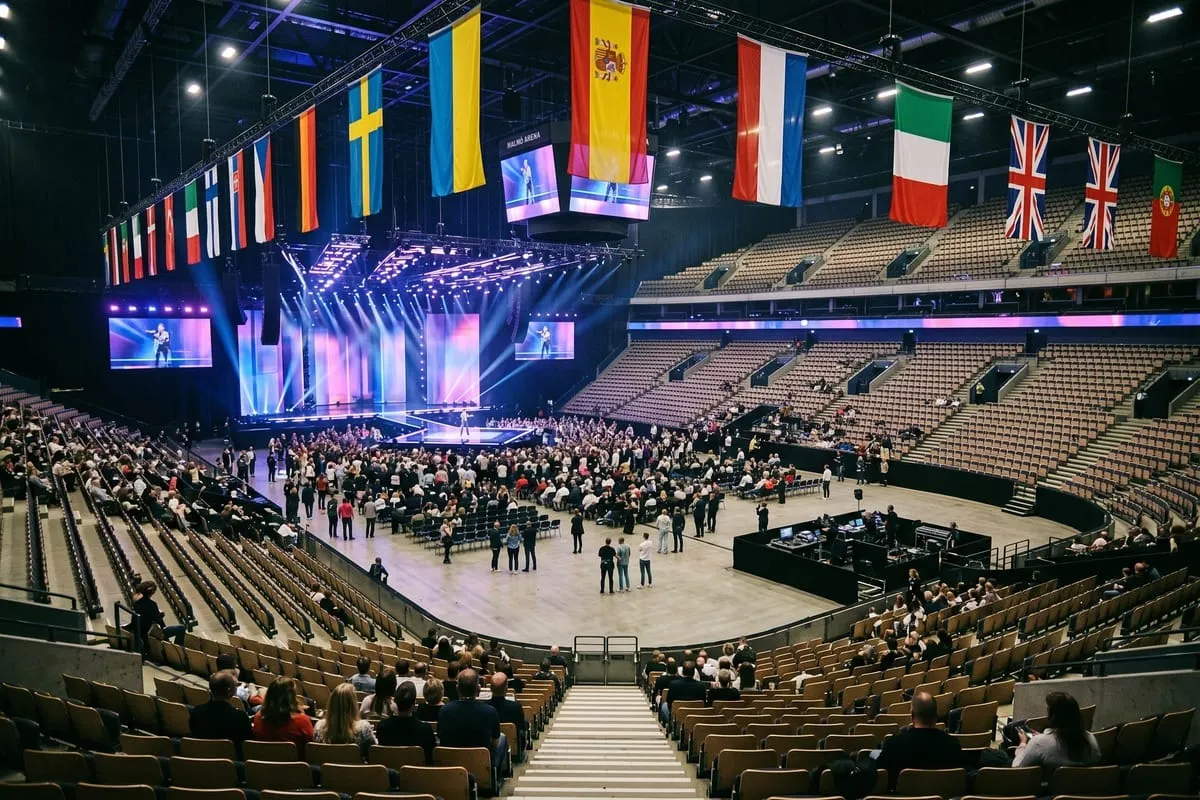 Grote arena met landenflags, verlicht podium, publiek op stoelen en open vloer tijdens songfestival-opbouw
