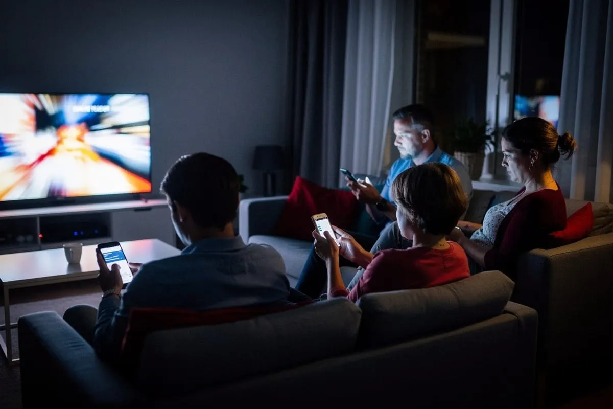 Familie op bank in donkere woonkamer, iedereen op smartphone terwijl tv aanstaat