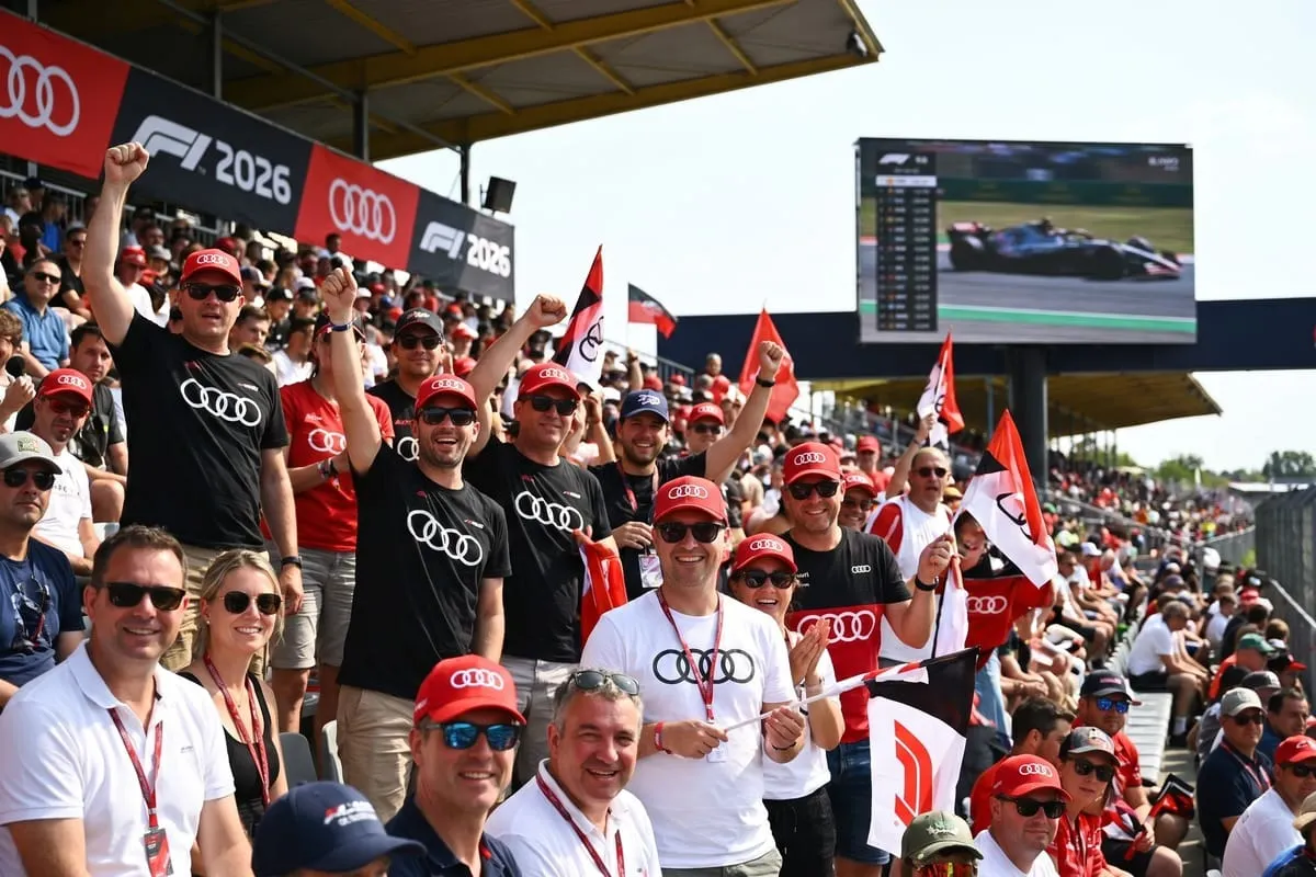 Groep supporters in zwarte Audi-shirts en rode petten juicht in tribune met vlaggen en F1 2026-borden