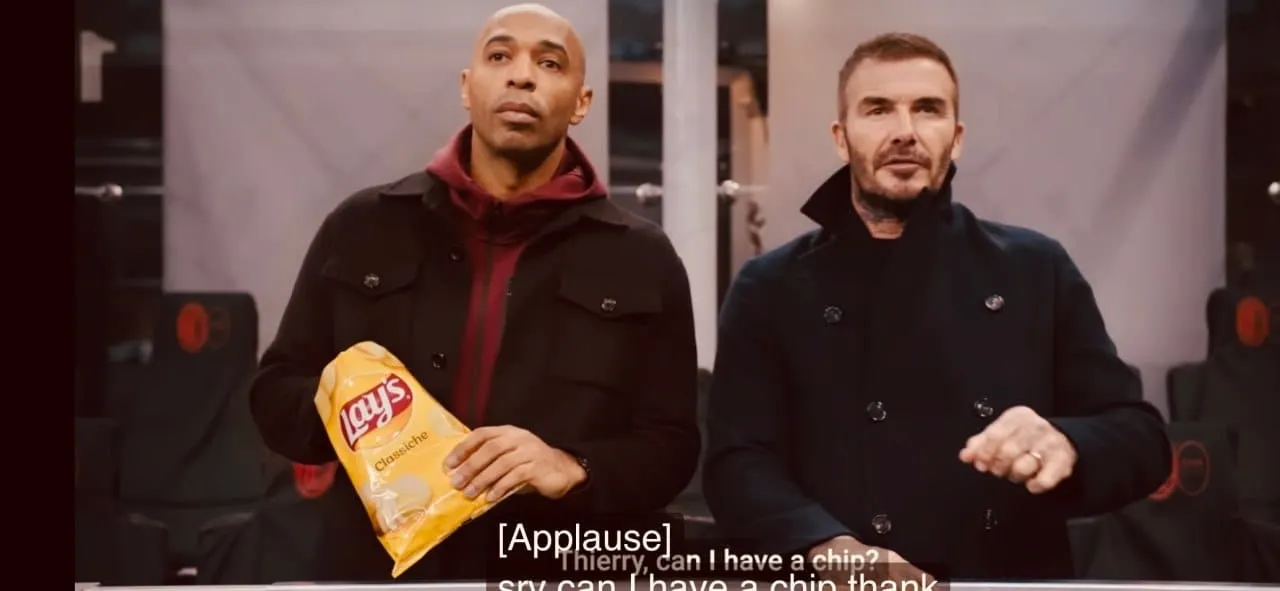Man in donkere jas houdt chipszak vast op straat met verlichte reclameborden en digitale schermen rondom