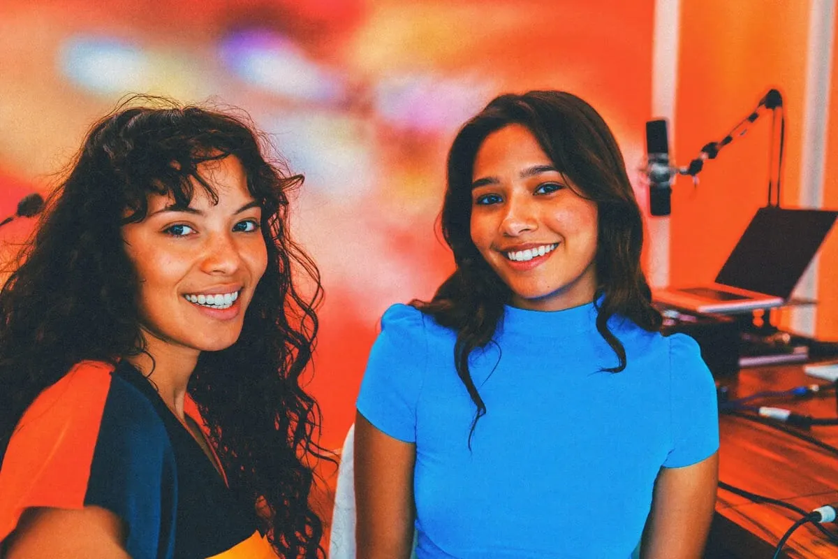 Twee lachende vrouwen in podcaststudio met microfoon en laptop, warme oranje verlichting