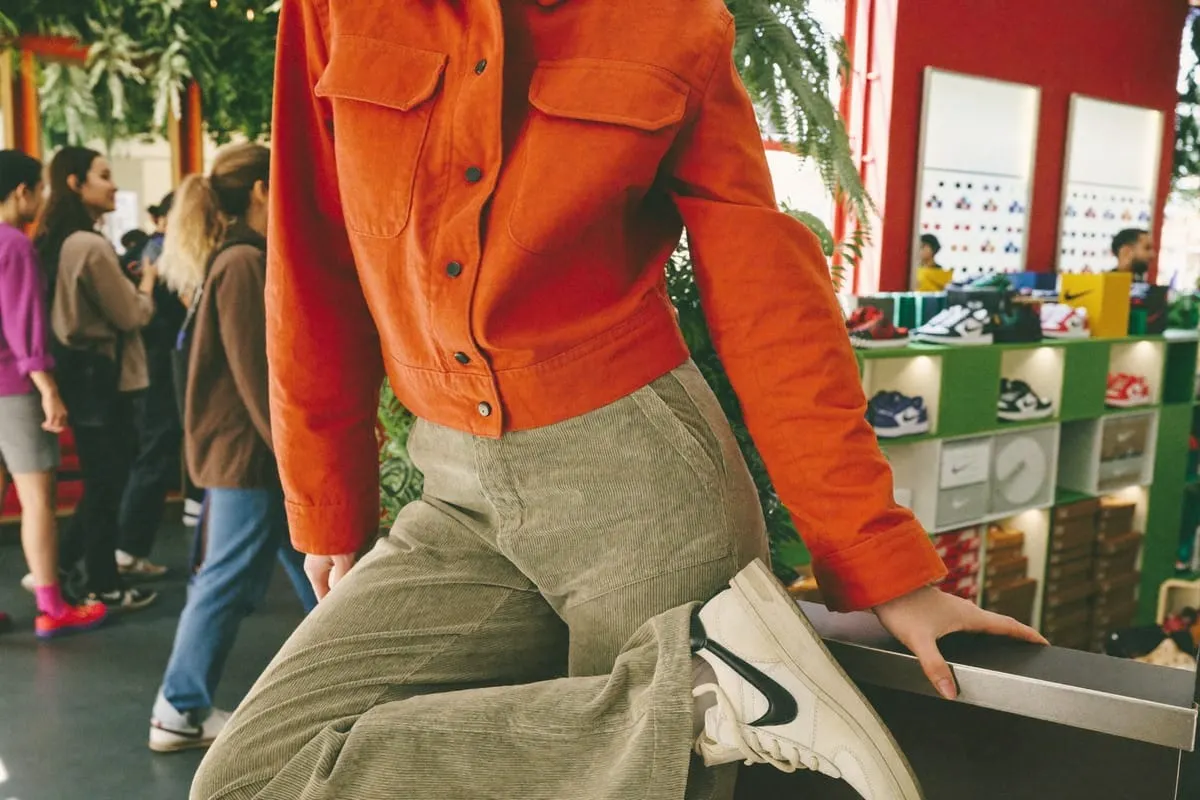 Persoon in oranje jack en corduroy broek zit op toonbank in sneakerwinkel met Nike-schoenen