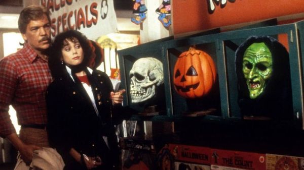 Tom Atkins and Stacey Nelkin stand behind three children wearing Silver Shamrock witch, skull, and pumpkin masks in Halloween III: Season of the Witch.
