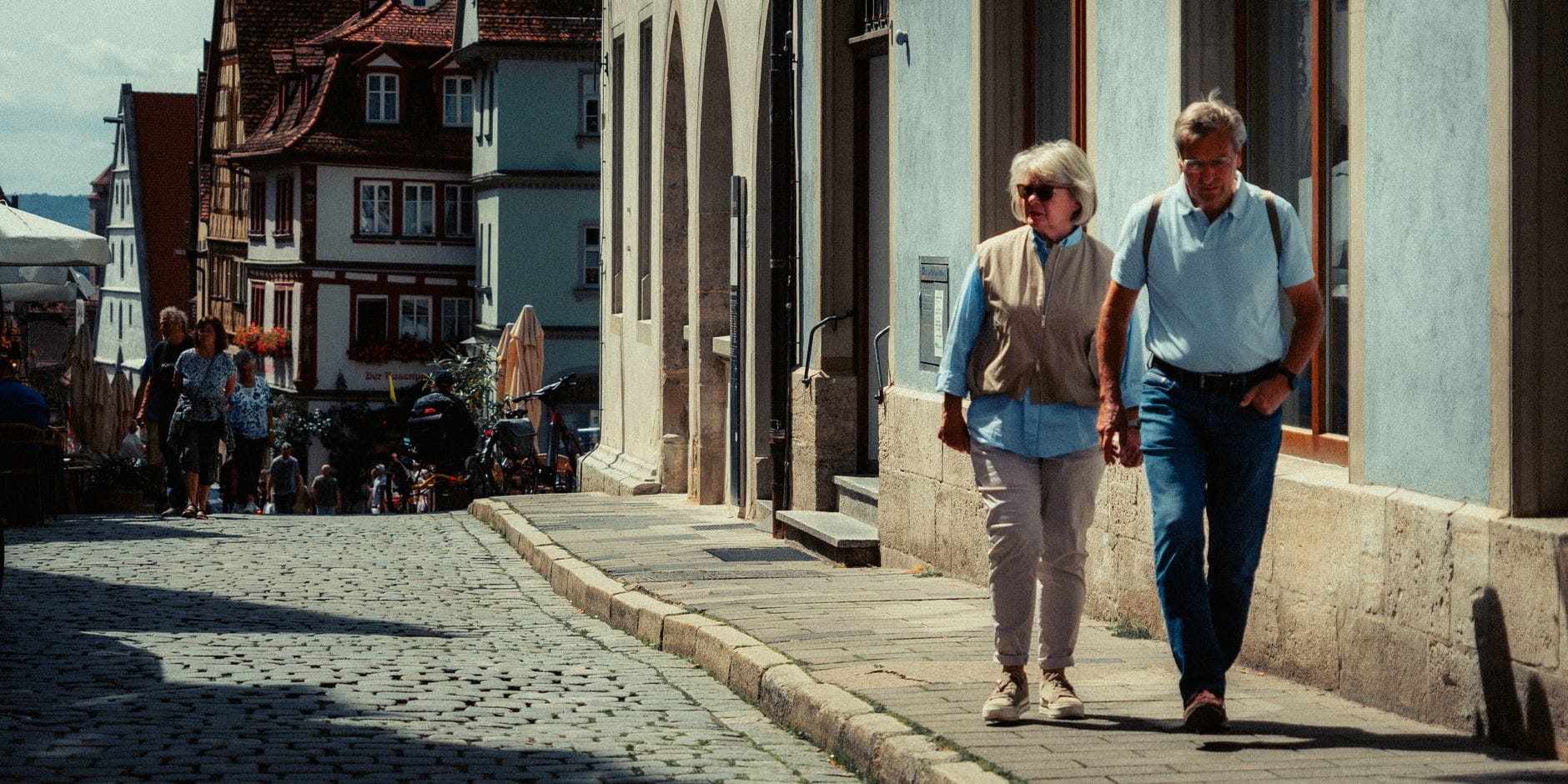 An elderly couple strolls along a cobblestone street in a charming European town, enjoying a sunny day.