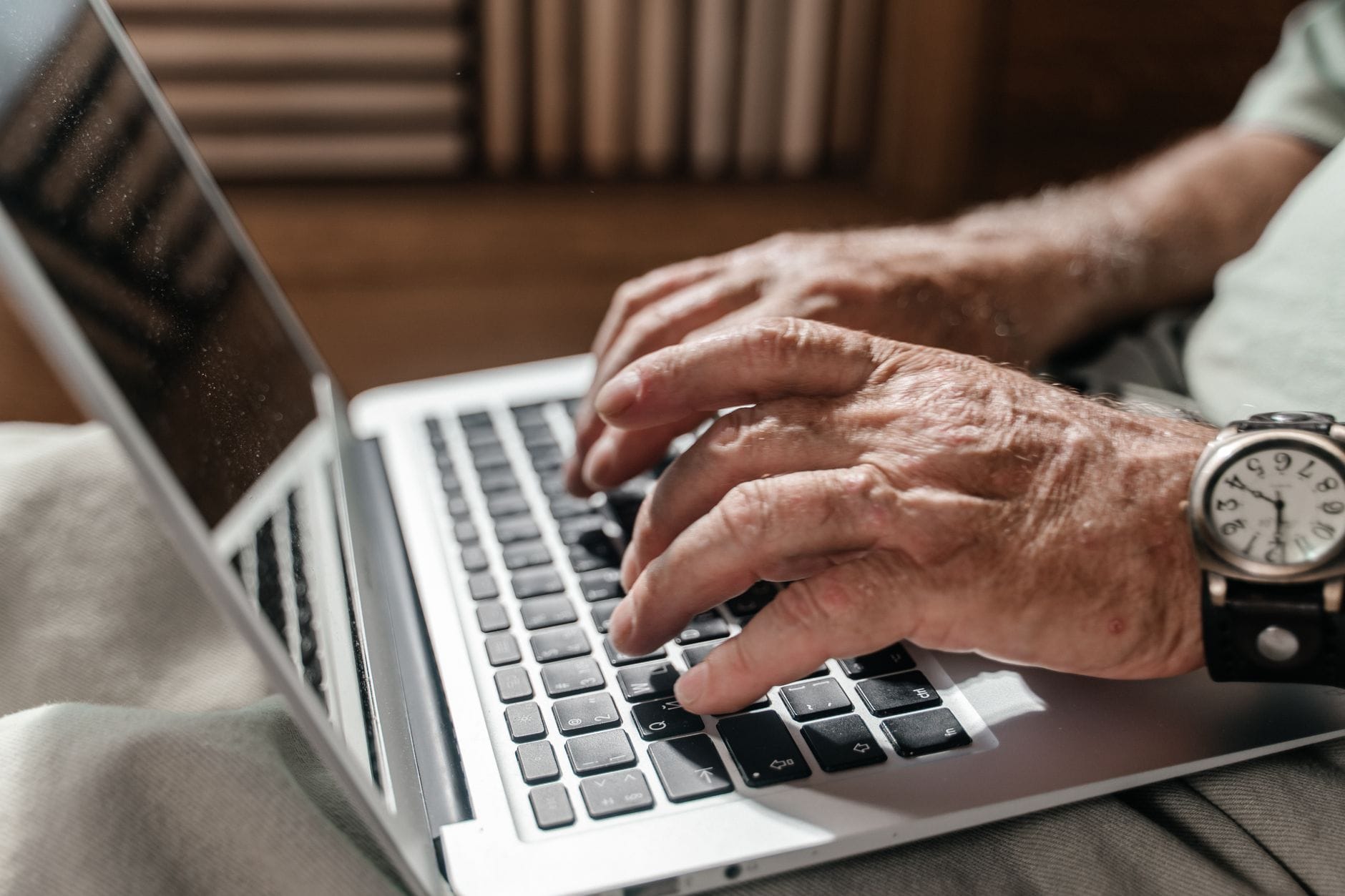Close-up of a senior adult's hands typing on a laptop, wearing a wristwatch, in a home office setup.