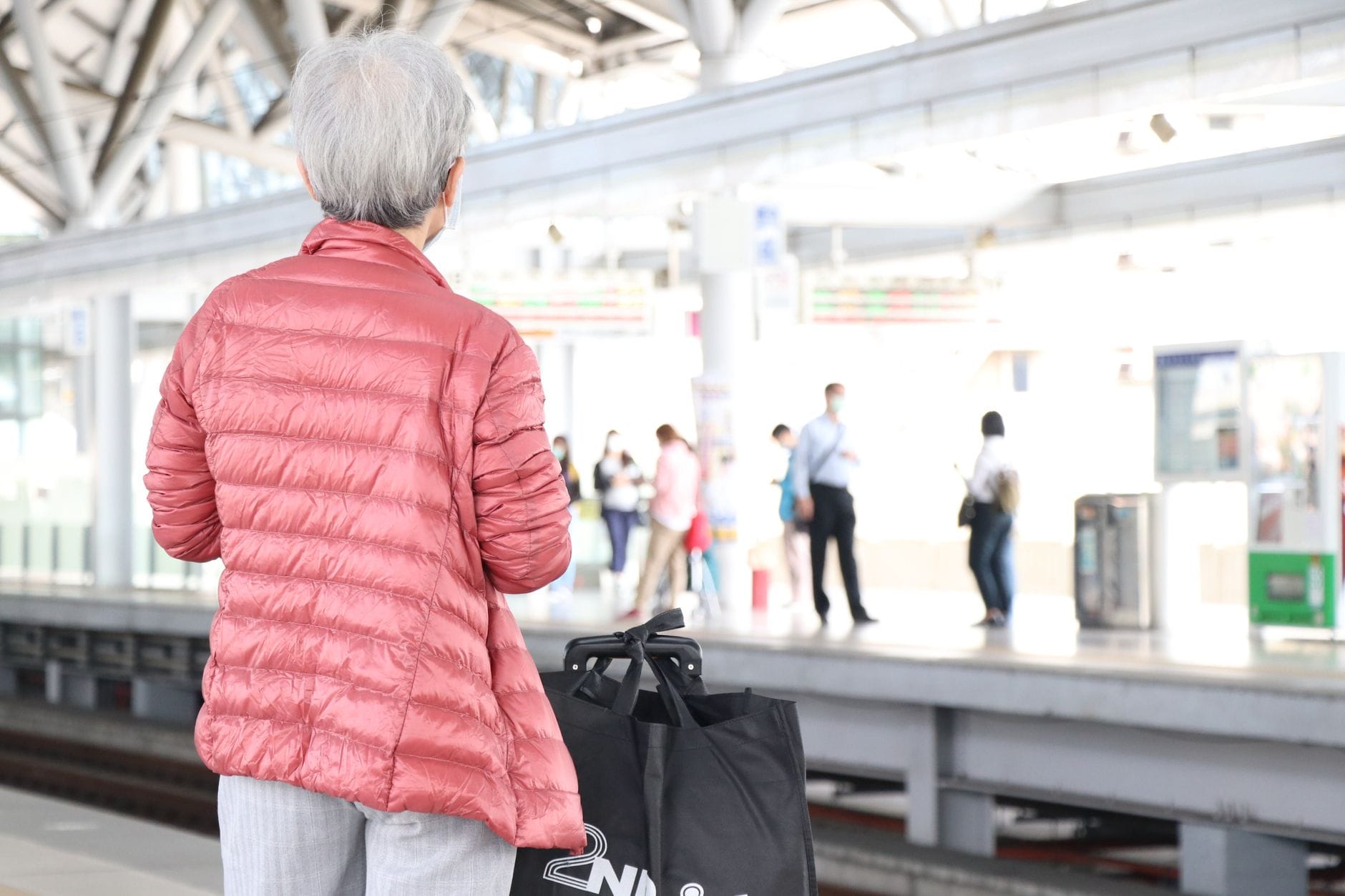 An elderly woman in a jacket waits at a bustling train station platform, holding luggage.