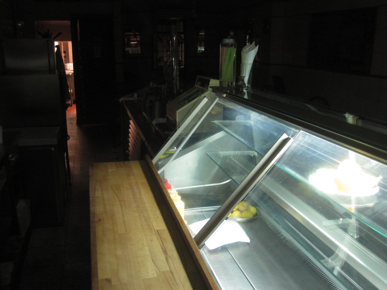 Behind the counter of an empty dark cafe at night. Lit only by the lights of the empty sandwich making station. 