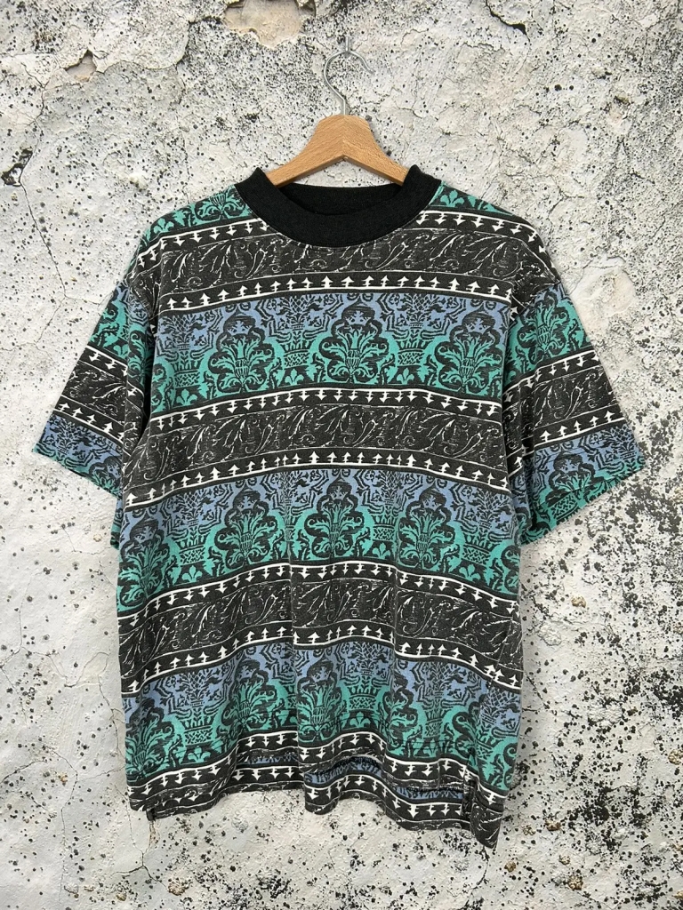 A very garish t-shirt on a hanger against a rough white wall. It has a teal and turquoise black and white pattern running horizontally. It's just horrid. 