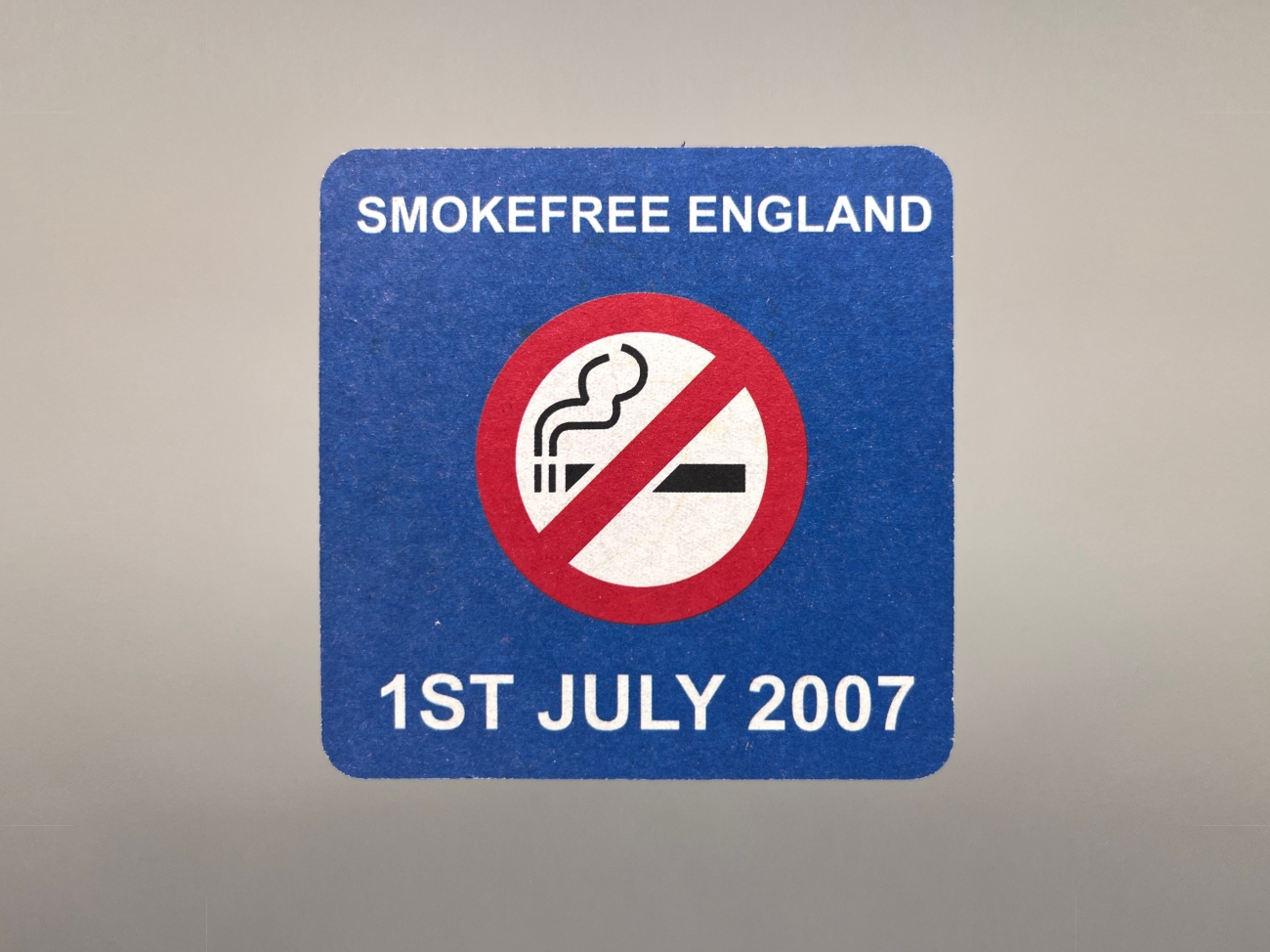 A blue beer mat with a no smoking sign in the middle. The top reads Smokefree England. At the bottom is says 1st July 2007