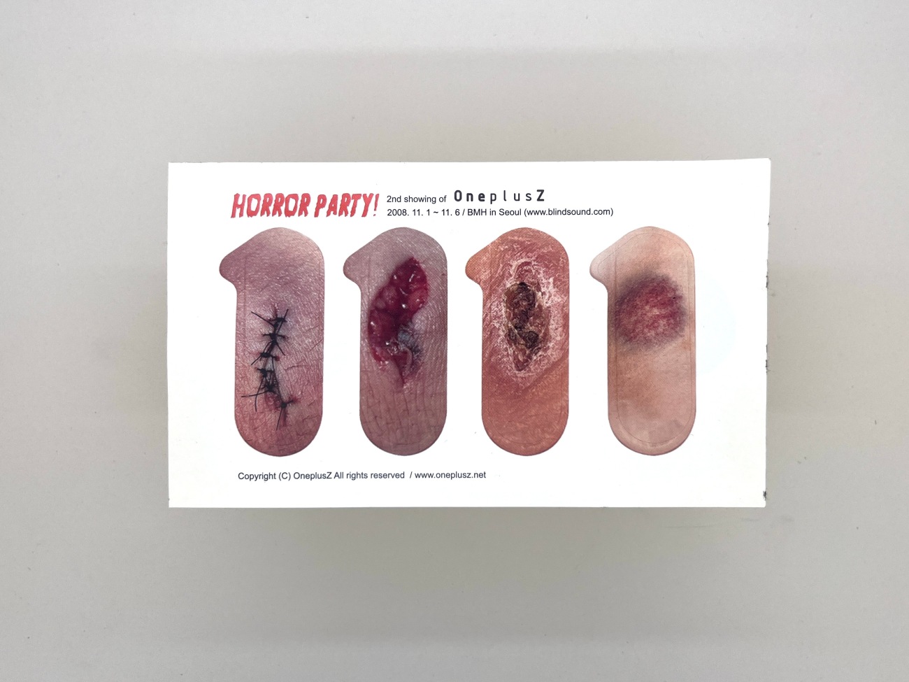 A postcard size piece of paper with the title Horror Party. It has 4 removable sticking plasters that have wounds printed on them. They're quite gross. One has stitches, one's a bruise and the other two are nasty cuts and scabs. They're really quite horrible. All also sadly on pale skin 