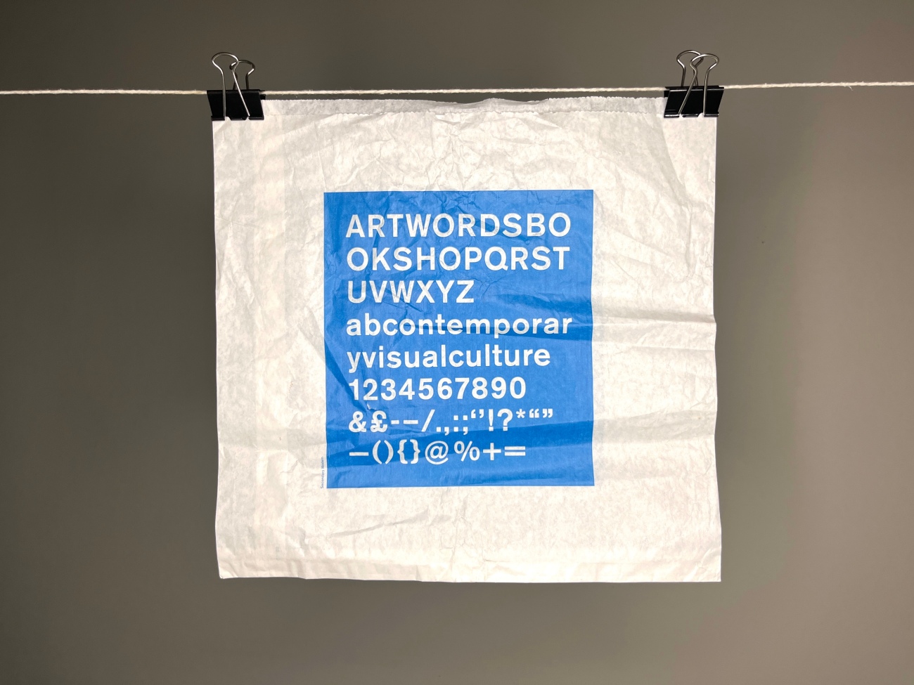 Hanging off a white string by two bulldog clips. A small white paper bag with a screen printed square of blue ink containing white letters. All caps with no spaces, it reads ART WORDS BOOK SHOP QRSTUVWXYZ and looks like it's just an alphabet. Under in lowercase with no spaces looking again just like a line of display of type it says ab contemporary visual culture 1234567890&£-–/.,:;''!?*""—{ }@%+=